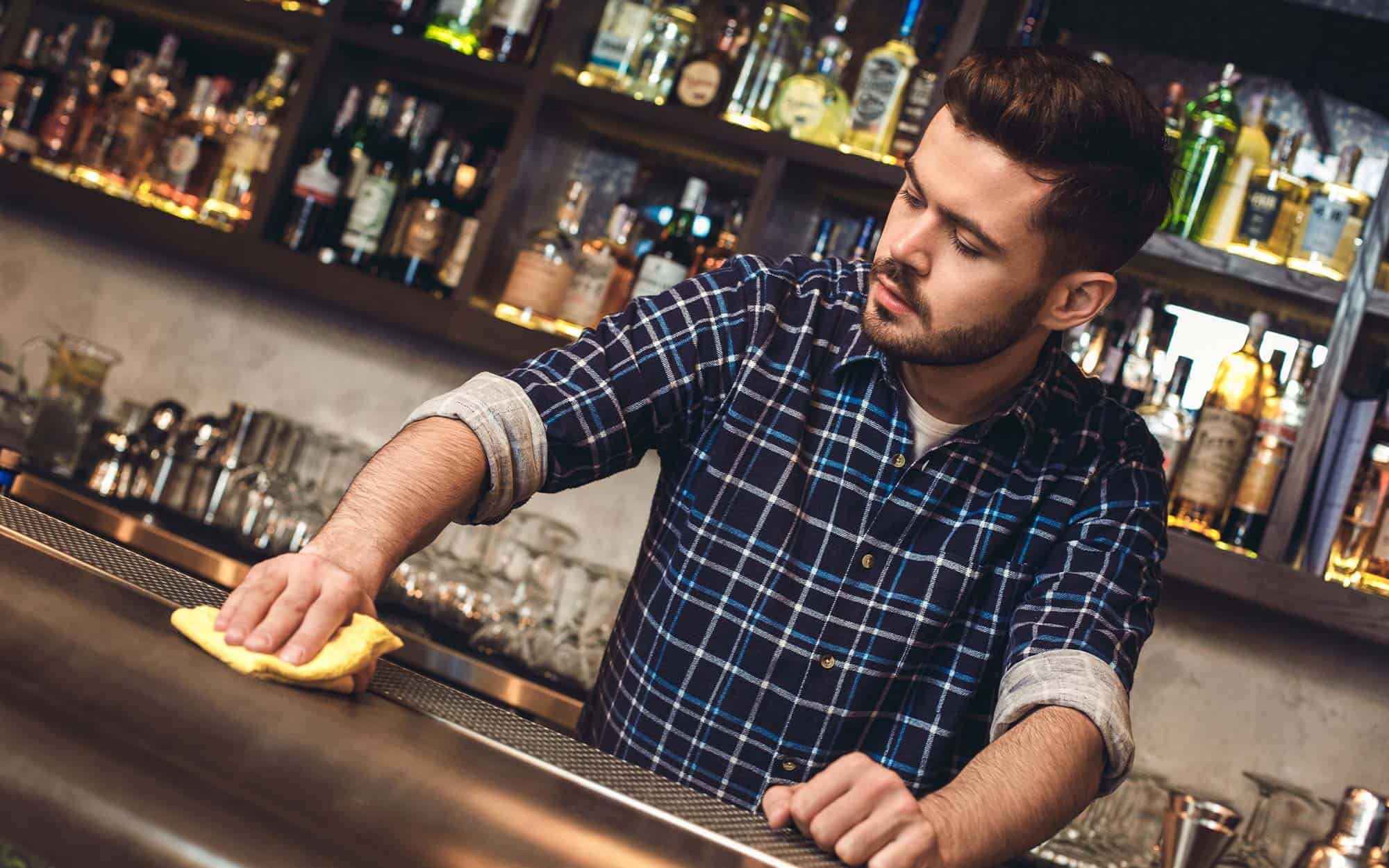 Young bartender standing at bar counter wiping surface concentrated